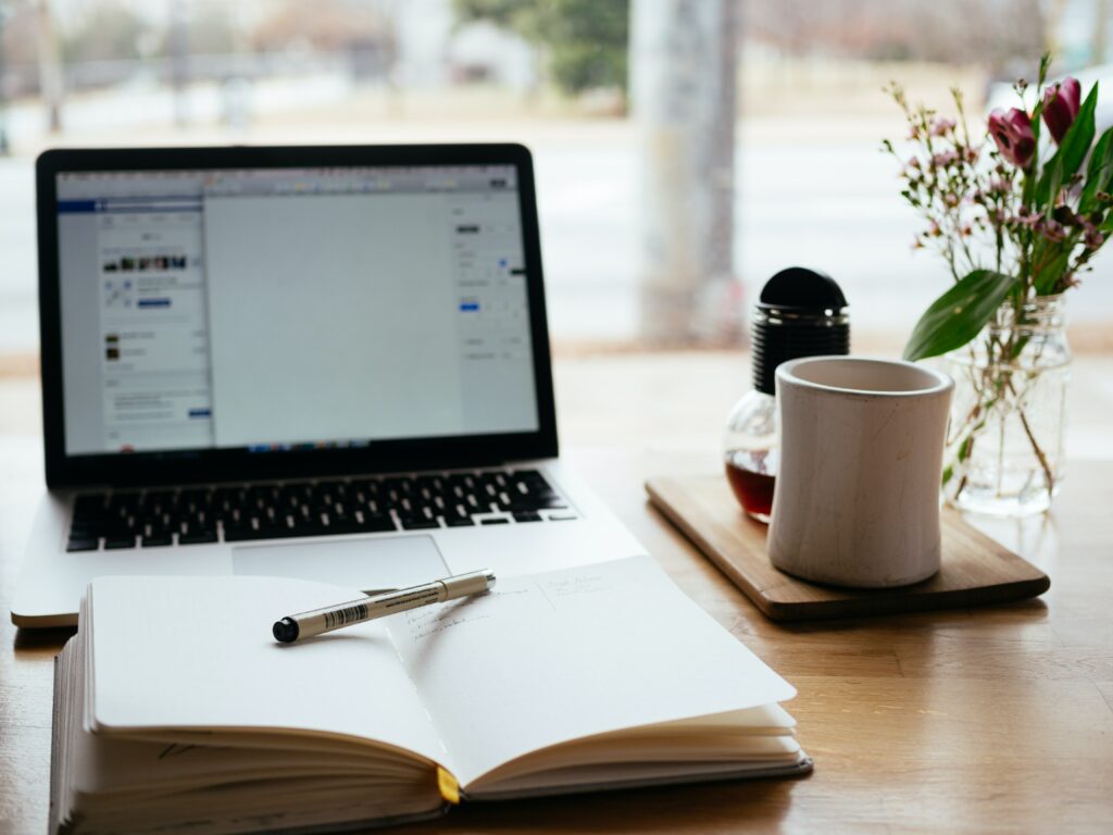 A desk with an open notebook with a pen on, a laptop with a document open and a coffee on a mat